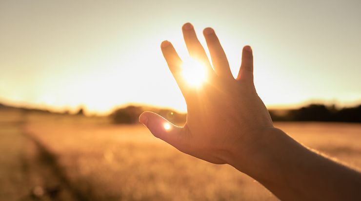 Blick auf eine untergehende Sonne über einem Feld, eine Hand verdeckt die Sonne teilweise