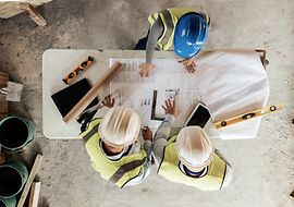 Vogelperspektive auf einen Tisch auf einer Baustelle, es stehen mehrere Baufachleute um den Tisch und besprechen sich